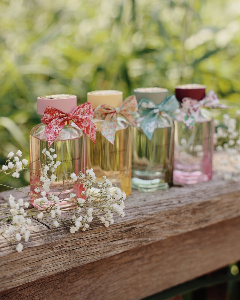 Four Laura Ashley parfum bottles with colourful bows on a wooden surface with blurred greenery in the background